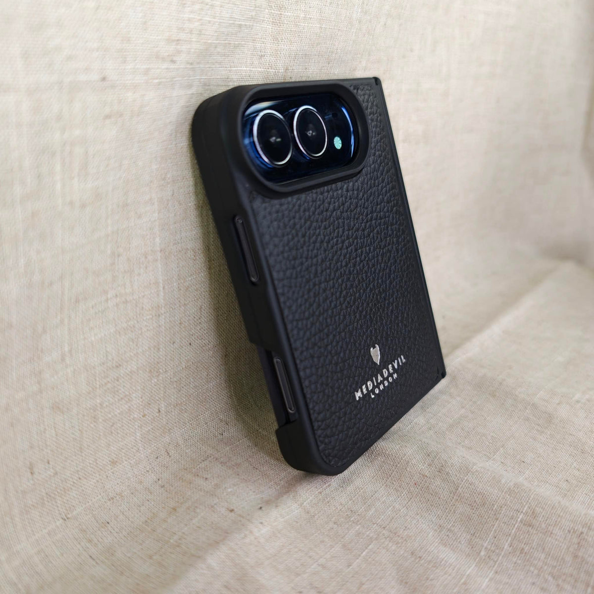 A black leather iPhone Duo phone case with a raised camera bar, shown standing upright on a beige linen surface. The case features a silver foil logo and text reading 'MediaDevil London' near the bottom.