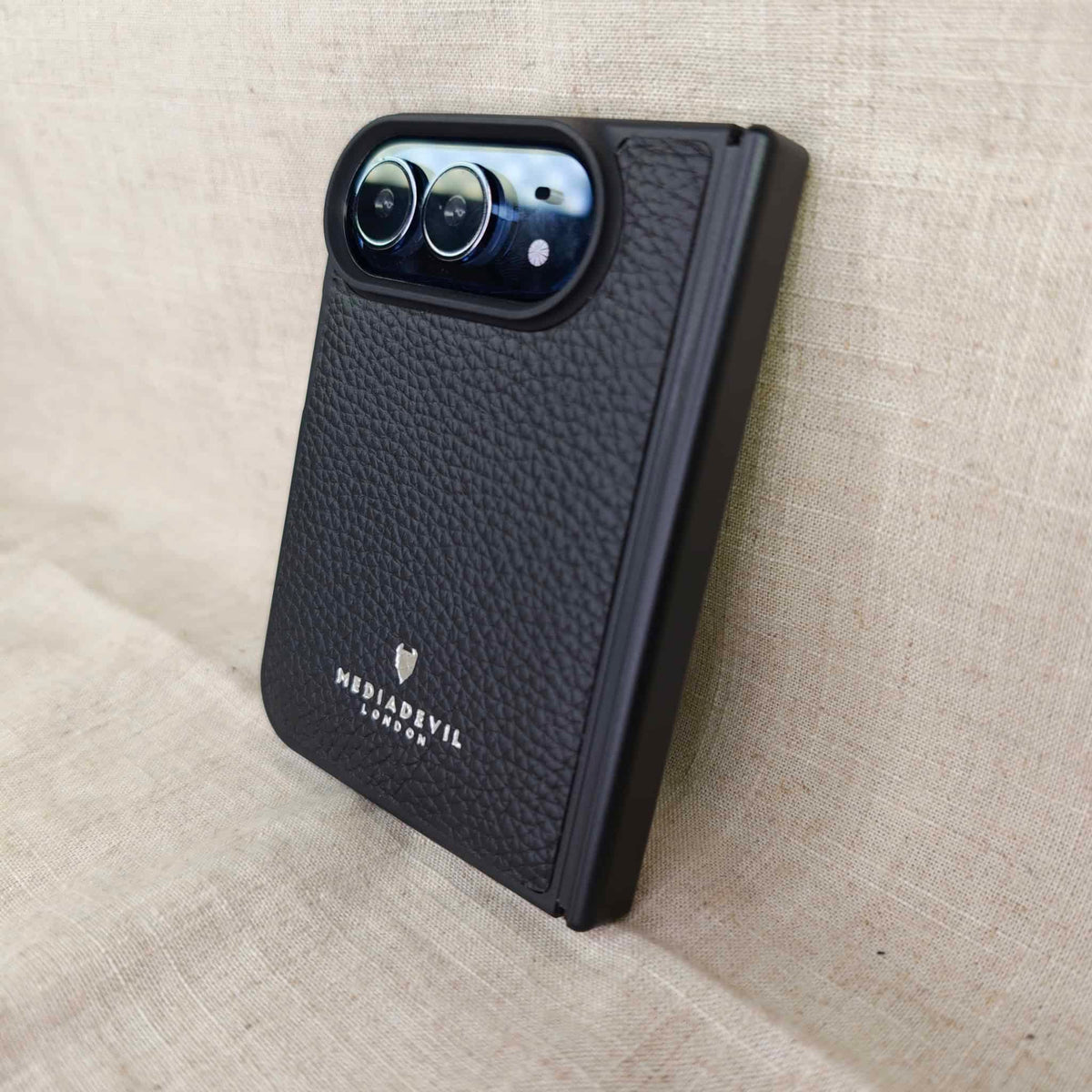 A black textured leather phone case with a raised camera bezel, featuring the 'MediaDevil London' logo, shown on a beige linen background.