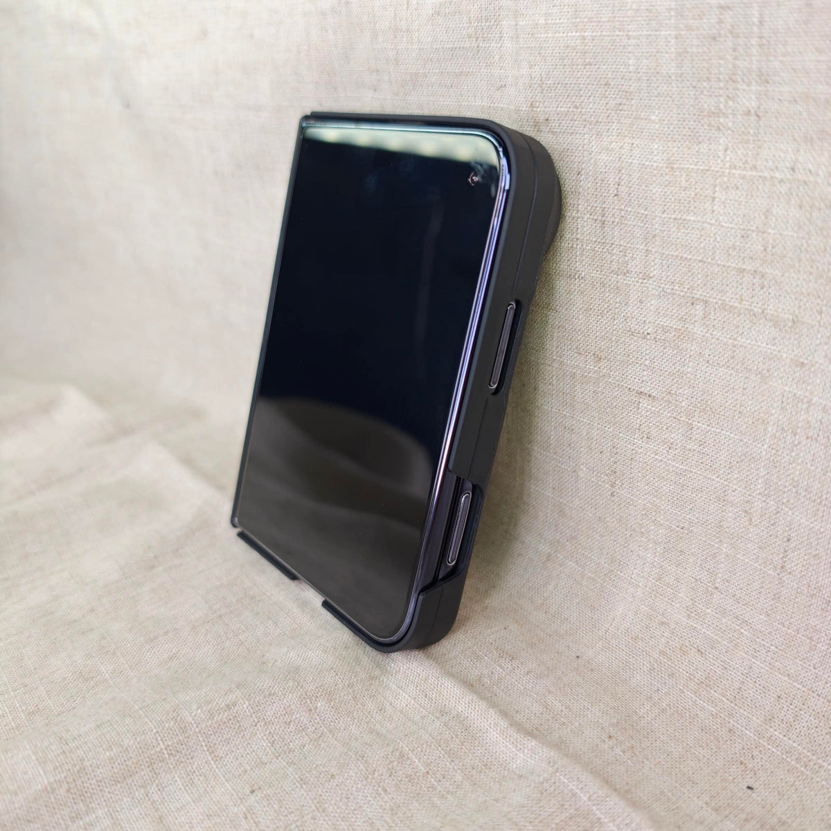 A black protective iphone duo case, shown standing upright on a textured beige fabric background.